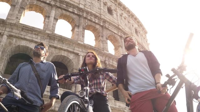 Three happy young friends tourists with bikes and backpacks at Colosseum in Rome having fun talking taking pictures on sunny day slow motion ground shot