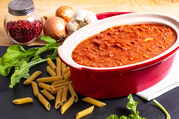 Homemade slow cooker bolognese sauce in red ceramic pot and raw penne, peppercorn, garlic, onion and rocket salad