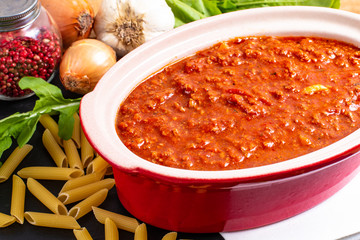 Homemade slow cooker bolognese sauce in red ceramic pot and raw penne, peppercorn, garlic, onion and rocket salad