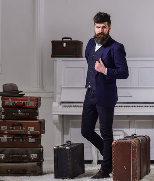 Macho stylish on thoughtful face standing near pile of vintage suitcase. Baggage and travelling concept. Man, traveller with beard and mustache with luggage, luxury white interior background.