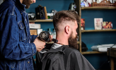 Barbershop concept. Hipster bearded client got hairstyle. Barber with hairdryer blows off hair out of cape. Barber with hairdryer works on hairstyle for bearded man, barbershop background