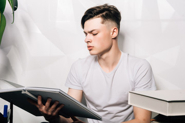 Cute young male writer romantically while working on new scientific book sitting at the desk with laptop and books on the gray vintage background.