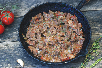 Stewed pieces of beef in an old frying pan and tomatoes, garlic, rosemary, pepper, on a rustic-style wooden background