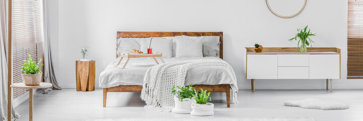 Panorama of a bright white and wooden bedroom interior with double bed and sideboard. Cushions, blanket, linen and food tray on the bed. Real photo. - obrazy, fototapety, plakaty