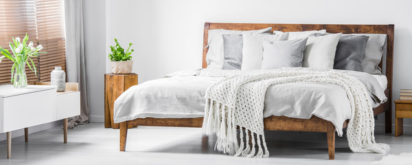 Wooden framed comfortable bed with many pillows, blanket and sheets and a sideboard with flowers on top in a white stylish bedroom interior. Real photo. Panorama. - obrazy, fototapety, plakaty