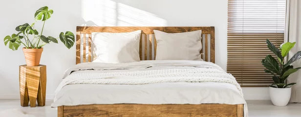 Foto op Aluminium Close-up of double wooden bed with bedding, pillows and blanket against white wall in a bright sunny bedroom interior. Two green plants standing beside. Panorama. Real photo. © Photographee.eu