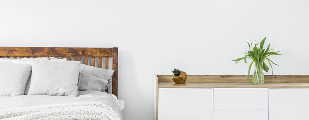 Close-up of a part of wooden bed with linen and pillows and a side cabinet with fresh cut flowers in a vase on top standing against a white wall in a bright bedroom interior. Panorama. Real photo.