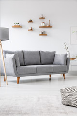 Grey couch in simple bright living room interior with pouf and lamp. Real photo