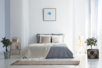Knit blanket on grey bed in bright bedroom interior with poster and plants. Real photo