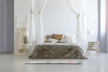 Lamp on table next to bed with pillows under canopy in bright scandi bedroom interior. Real photo