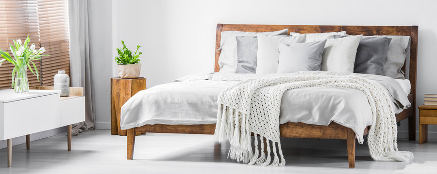Wooden framed comfortable bed with many pillows, blanket and sheets and a sideboard with flowers on top in a white stylish bedroom interior. Real photo. Panorama.