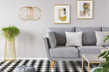Real photo of a living room interior with gold posters on the walls, grey sofa with cushions and checkered floor