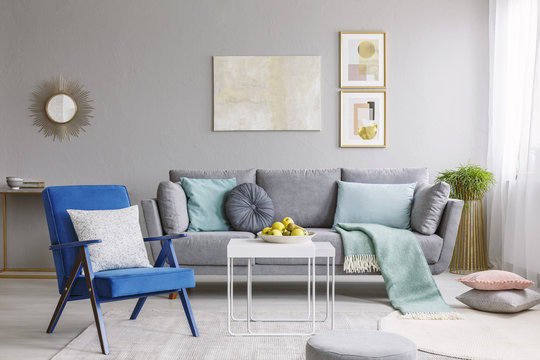 Real photo of a blue armchair standing next to a white table in a modern living room interior with a grey sofa and posters