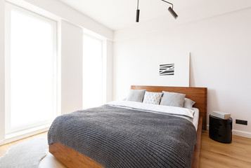 Grey blanket on wooden bed in white bright bedroom interior with simple poster. Real photo