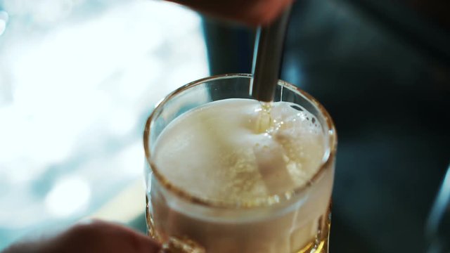 Young employee is filling glass with beer in trendy bar. Alcoholic beverage is poured into mug from tap equipment indoors. Light foamy drink pours with thin stream into transparent glass from steel