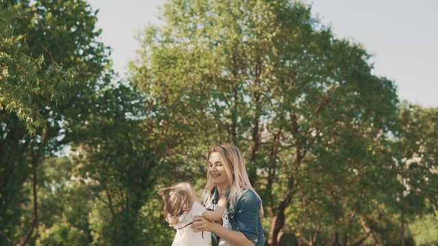 Loving caucasian mother in dress with her little daughter having fun and playing in park. Happy mom spinning her laughing daughter in slow motion