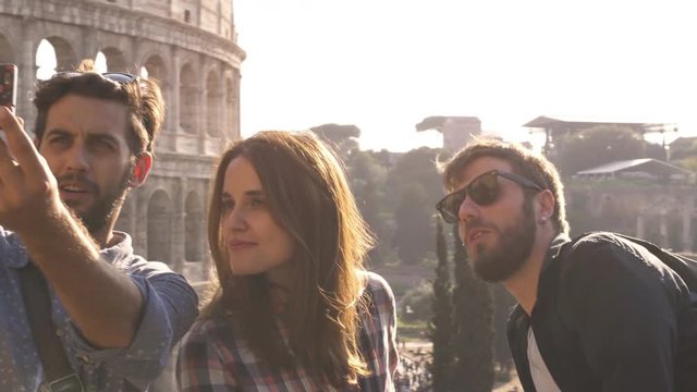 Three happy young friends tourists at Colosseum in Rome taking funny selfies with smartphone stick making faces on hill at sunset with trees slow motion steadycam