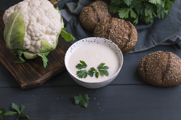 Cream of cauliflower soup with cream and greens, in a white cup on a wooden board and black background. Vintage photo. Fresh greens, cauliflower and grain bread. Healthy fitness dinner. Copy space,