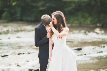 Fototapeta na wymiar Elegant stylish happy brunette bride and gorgeous groom on the background of a beautiful river in the mountains