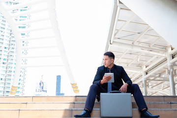 Portrait charming handsome young businessman. Attractive handsome business man is sitting on step and looking at copy space. He is holding tablet and thinking his work in summer time. city background