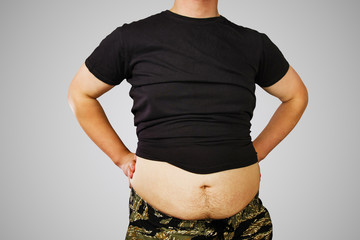 Torso of young fat guy in gray t-shirt holds his hands on belt, on gray background.