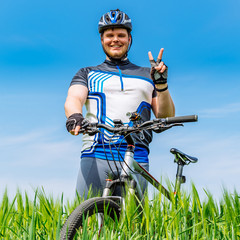 young adult smiling man standing with bicycle in green barley fi