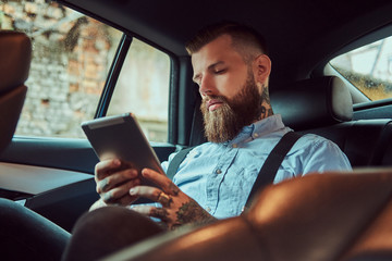Old-fashioned tattooed hipster guy in a shirt with suspenders, using a tablet while sitting in a luxury car on back seat.