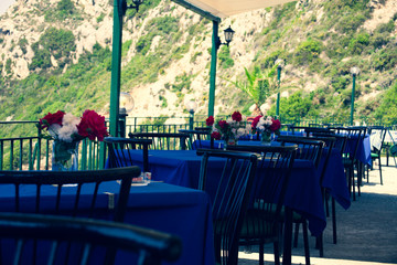 Restaurant tables with black chairs red and white flowers and blue tablecloth in a natural environment. Peaceful place to have food. Having meal on table with great view