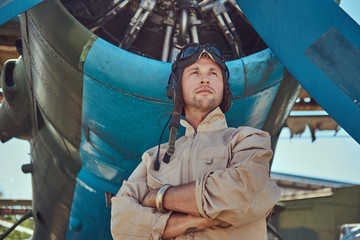 Valiant handsome pilot in a full flight gear standing with crossed arms near military airplane. © Fxquadro