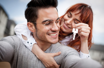 Loving couple with keys to their new home hugging and looking at camera taking selfie