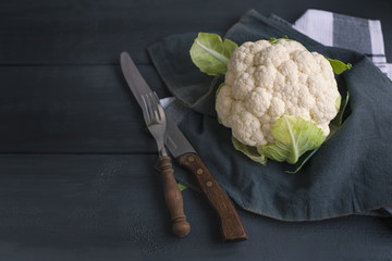 Cauliflower on a black wooden table, old fork, knife and a kitchen towel. Free space for text.
