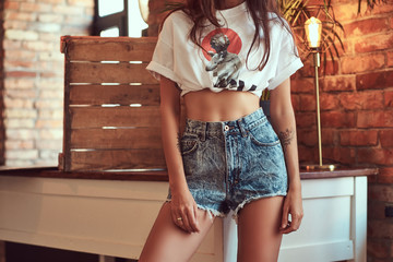 Cropped image of a sexy slim cool girl in a white top and denim shorts, standing near a table in...