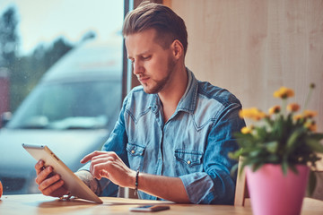 Hipster with a stylish haircut and beard sits at a table in a roadside cafe, looks something in the tablet.