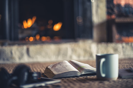Shallow depth of field photo of open Bible, coffee, notepad and tablet in front of fire place