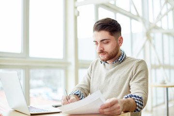 Young man working with papers and watching laptop while writing at table in light room