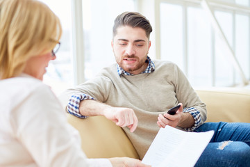Obraz premium Young man with smartphone sitting on couch with woman reading papers and doing business. 