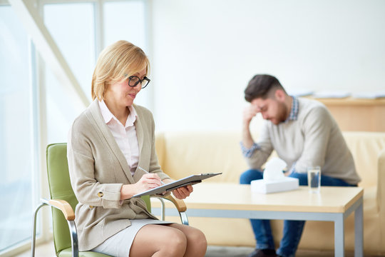 Elegant woman taking notes on clipboard while conducting therapy with upset man having mental problems