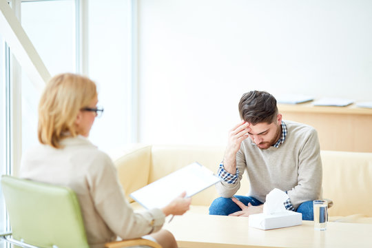 Woman with clipboard consulting young man having problems after emigration sitting in light cabinet