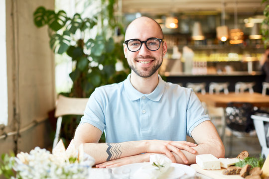Handsome bald young male in glasses sitting with crossed hands and smiling on background of blurred restaurant