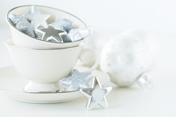 Photograph of White teacups filled with silver stars and silver Christmas ornaments on white with white Christmas ornaments 