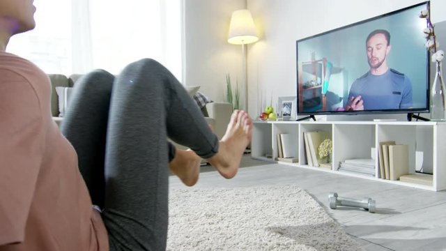 PAN of male online personal coach instructing young woman in sportswear doing knee tuck crunches on floor, then letting her rest