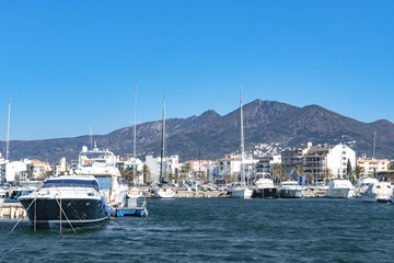 Fototapeta na wymiar Motor and sailboats ancoring in the marina in Roses, Spain. The water is colored deep blue, while the sky has a lighter tint. In the back the start of the pyrenees can be seen