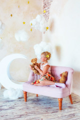 Blonde teen girl plays with teddy bear sitting on pink couch in background of large moon and clouds in room