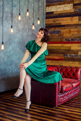 Smiling girl in green dress sitting on edge of red stylish sofa on background of wooden wall