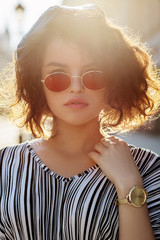 Outdoor close up portrait of young beautiful stylish curly girl wearing oval red color sunglasses,...