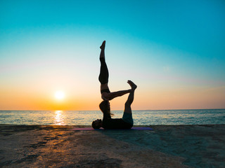 Silhouettes of fit young couple doing acro-yoga at sea beach. Man lying on concrete plates and balancing woman in his feet. Beautiful couple practicing yoga together