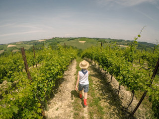Cute little boy walking in vineyard between the rows in a sunny summer day. A kid with straw hat and red boots treading trough the path on the Italian hills in Monferrato. Blue sky over the hills