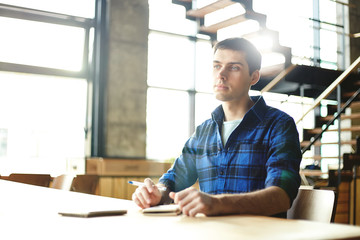 Casual young man looking away contemplating on idea while holding pen and sitting with notebook at desk. .