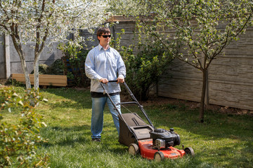 Young man in sunglasses, jeans and shirt mows the grass on the lawn in the village in the summer.