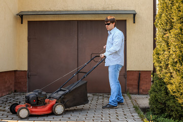 Young man in sunglasses starts a lawn mower opposite the garage in the summer.